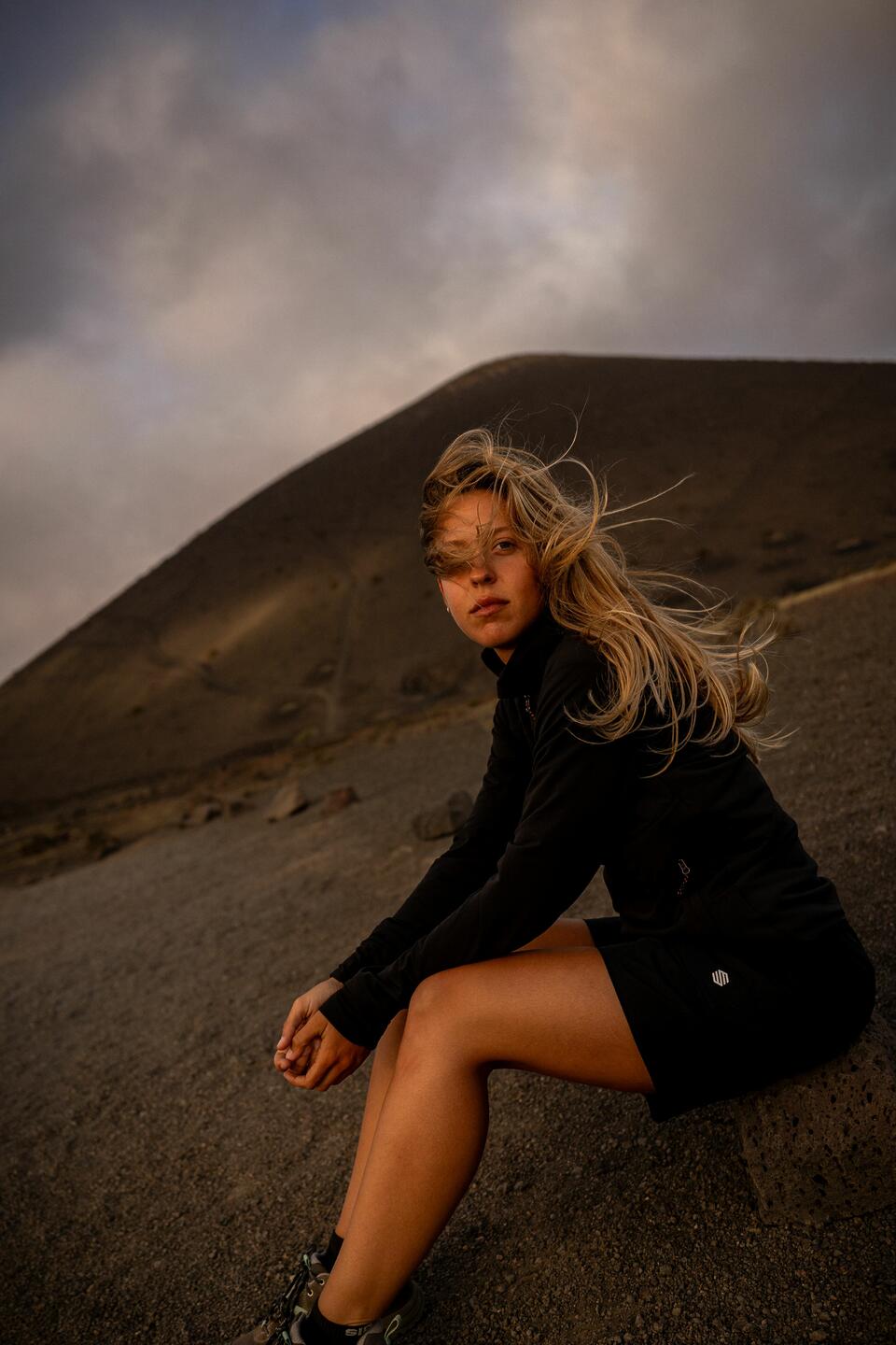 A seated blonde woman, dressed in black Siroko Adventure clothing, with a rocky mountain behind her.